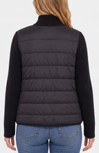 Back view of a person wearing a black quilted vest over a black ribbed sweater and blue jeans.
