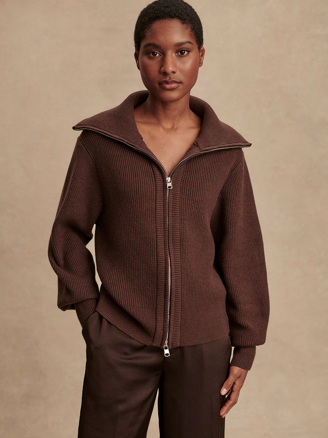 Woman wearing a brown ribbed knit zip-up jacket with wide collar and matching brown pants against a beige background