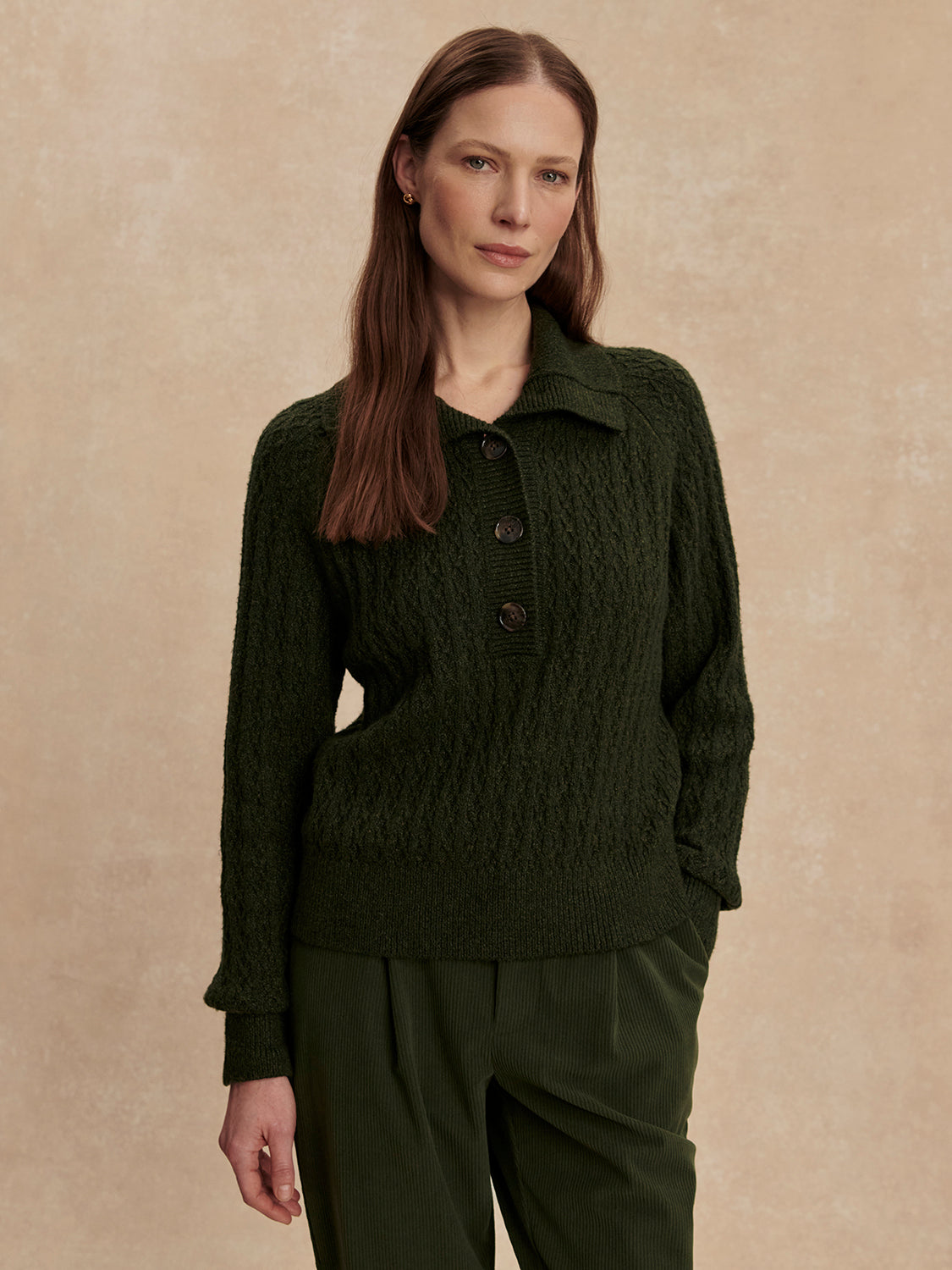 Woman with long brown hair wearing a dark olive Bridget half button sweater and matching dark olive pants standing against a beige background