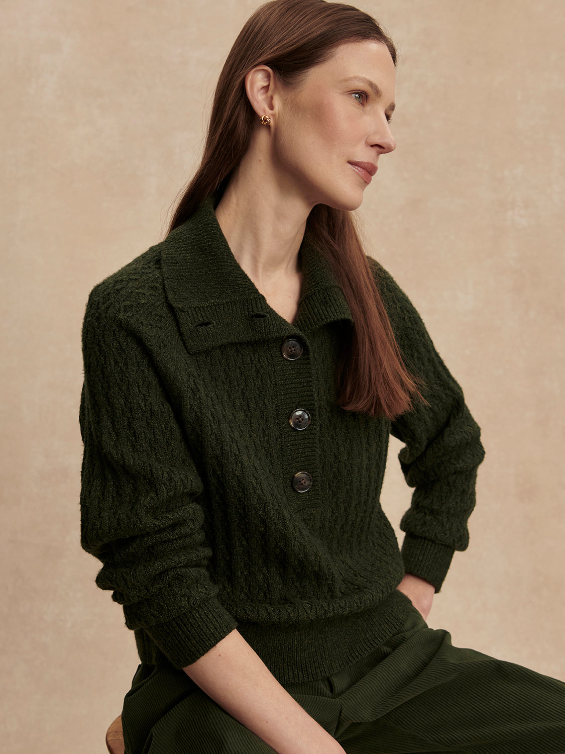 Woman with long brown hair wearing a dark olive half button cable knit sweater sitting against a beige backdrop