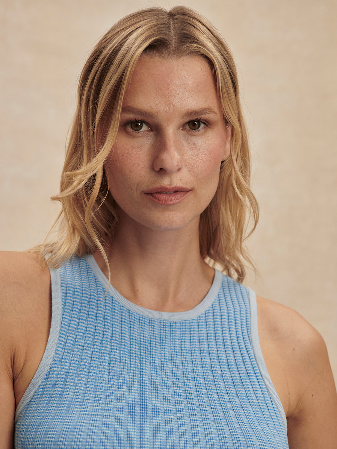 Close-up of a woman wearing a Mist Blue Hanson Rib Knit Tank by Varley with soft waves, neutral makeup, and a beige background.