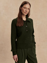 Woman with long straight brown hair wearing a dark olive half button textured sweater and matching pants standing against beige wall