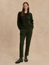 Woman with straight brown hair wearing a dark olive half button sweater and matching pants standing against a beige background