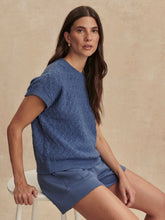 Woman wearing a blue knitted top and shorts sitting on a white stool against a beige background