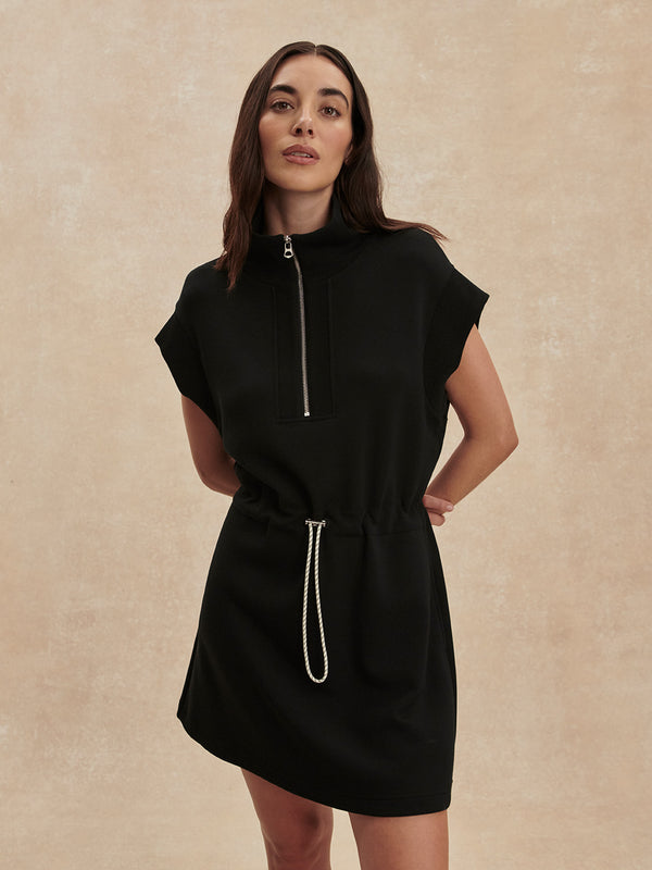 Woman wearing a sleek Black Avaya Half Zip Dress by Varley with a drawstring waist and short sleeves.