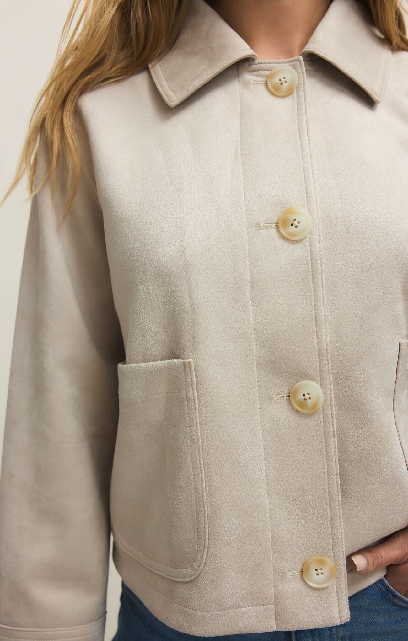 Close-up of woman wearing a light beige suede jacket with large buttons and patch pockets over blue jeans